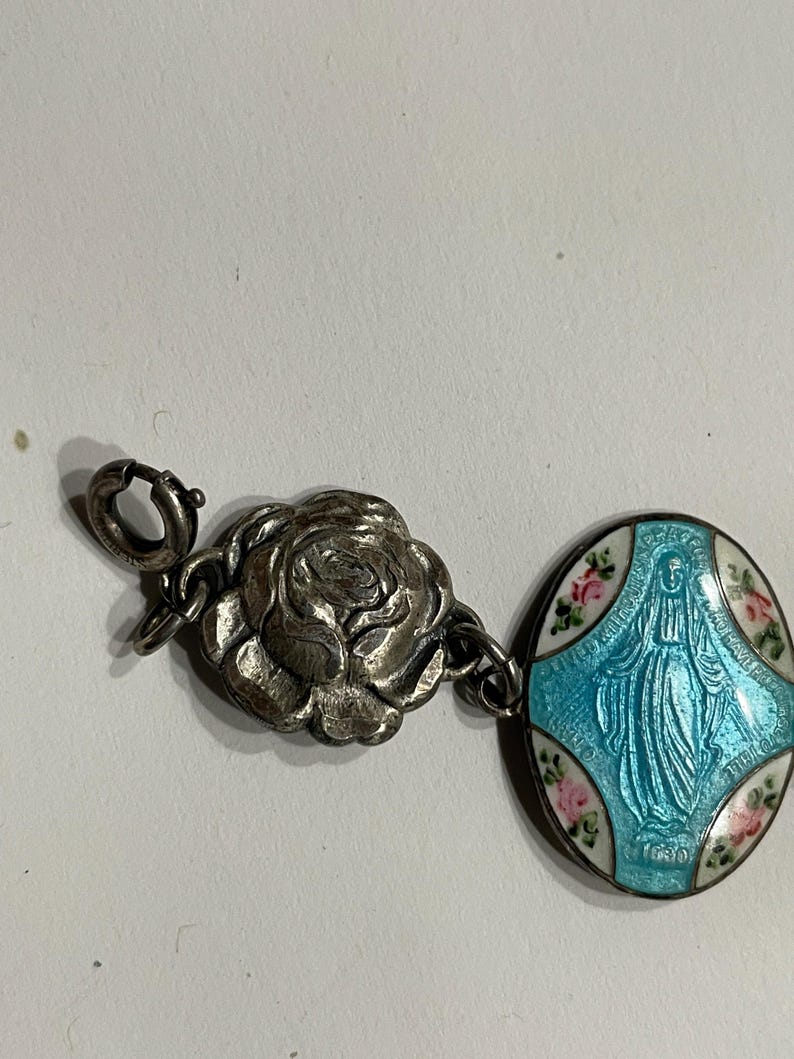 May include: A silver-toned charm bracelet component featuring a three-dimensional rose charm connected to an oval pendant. The pendant displays a blue enameled figure of the Virgin Mary surrounded by white enamel with small pink floral accents.