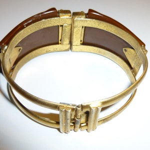 May include: A gold-toned bangle bracelet with a brown plastic insert. The bracelet is hinged and has a double band design.