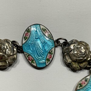 May include: A bracelet featuring oval-shaped charms with a light blue enamel depiction of a figure in robes, surrounded by white enamel and small pink and green floral accents. The charms are connected by metallic silver-toned rose-shaped links.
