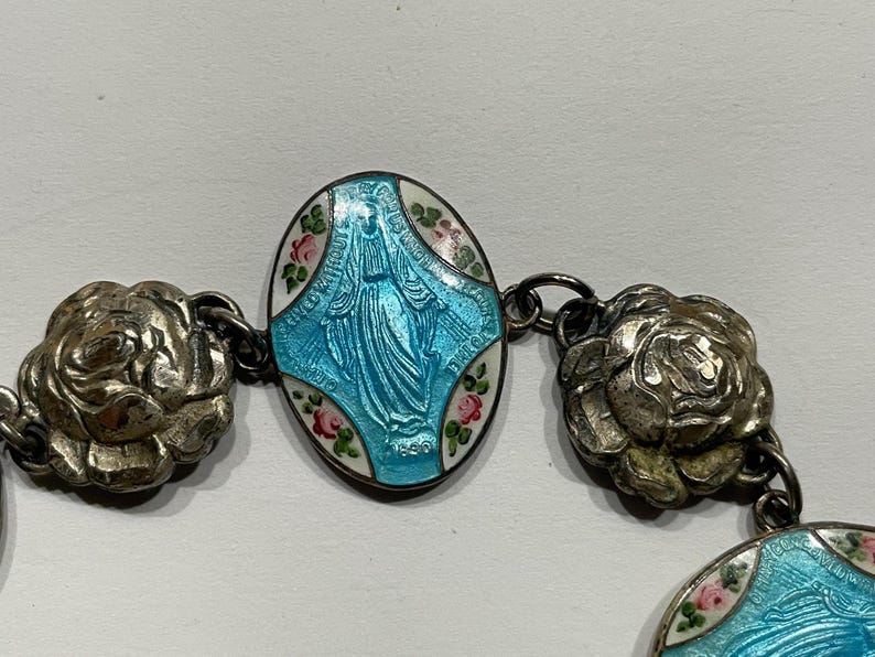 May include: A bracelet featuring oval-shaped charms with a light blue enamel depiction of a figure in robes, surrounded by white enamel and small pink and green floral accents. The charms are connected by metallic silver-toned rose-shaped links.