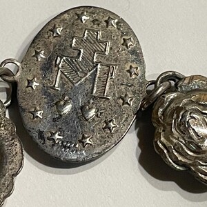 May include: A silver-toned metal bracelet featuring an oval charm embossed with a cross, the letter M, two hearts, and a border of stars. The bracelet includes additional circular charms, one depicting a rose and another showing a profile portrait.
