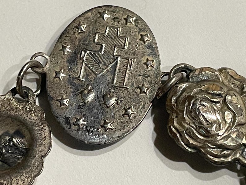 May include: A silver-toned metal bracelet featuring an oval charm embossed with a cross, the letter M, two hearts, and a border of stars. The bracelet includes additional circular charms, one depicting a rose and another showing a profile portrait.