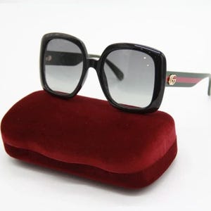 May include: Black square-framed sunglasses with gradient lenses rest on a burgundy velvet case. The sunglasses feature a green and red stripe on the temples and a gold logo. The case is rectangular and has a rounded top.
