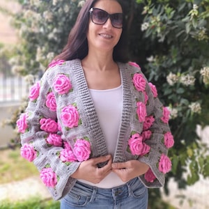 May include: A gray crocheted cardigan with pink 3D flower embellishments. The cardigan has a V-neck and long sleeves.