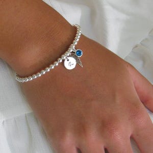 May include: A silver beaded stretch bracelet featuring a small round charm engraved with the letter R, a delicate silver feather charm, and a single round blue crystal charm. The bracelet is composed of numerous small, polished silver spheres.