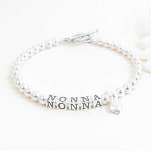 Puede incluir: Pulsera de cuentas plateadas con un dije de corazón y la palabra "NONNA" escrita en cuentas plateadas.