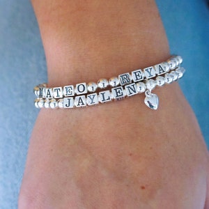 Puede incluir: Dos pulseras de cuentas plateadas con los nombres "Mateo", "Jaylen" y "Reya" escritos en letras negras. Un pequeño dije de corazón plateado cuelga de la parte inferior de las pulseras.