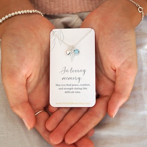 May include: A silver necklace featuring a small heart charm and a blue crystal charm, suspended from a delicate silver chain. The necklace is presented on a white card with the inscription "In loving memory" and a message offering peace, comfort, and strength. The card also includes the website address "www.PickledBeads.Etsy.com".