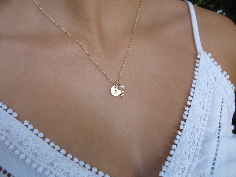 May include: A gold necklace with a small round pendant featuring the letter "S" and a pearl charm. The necklace is worn by a person with a white lace top.