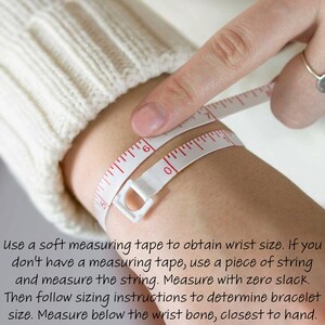 May include: A close-up of a person's wrist being measured with a white measuring tape. The tape is wrapped around the wrist and the person is holding the end of the tape with their finger. The text on the image reads: "Use a soft measuring tape to obtain wrist size. If you don't have a measuring tape, use a piece of string and measure the string. Measure with zero slack. Then follow sizing instructions to determine bracelet size. Measure below the wrist bone, closest to hand."