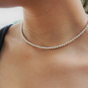 May include: A silver beaded choker necklace on a person's neck. The necklace is made of small, round beads and is worn close to the neck.