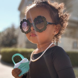 May include: A young girl wearing a black dress, a pearl necklace, and large black sunglasses with flower-shaped frames. She is holding a small turquoise handbag with the text "Dior & Co. Est. 2020" printed on it.