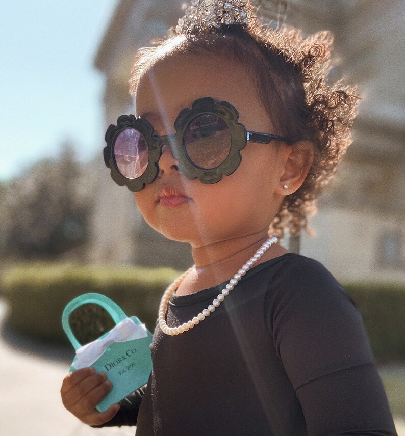 May include: A young girl wearing a black dress, a pearl necklace, and large black sunglasses with flower-shaped frames. She is holding a small turquoise handbag with the text "Dior & Co. Est. 2020" printed on it.