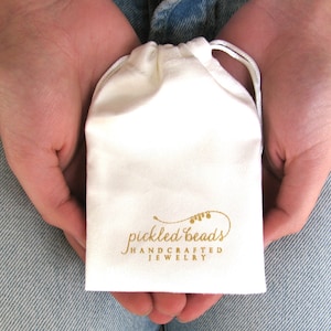May include: A small, off-white drawstring pouch with the words "pickled beads HANDCRAFTED JEWELRY" printed in gold. The pouch is held in open hands, suggesting it contains jewelry.