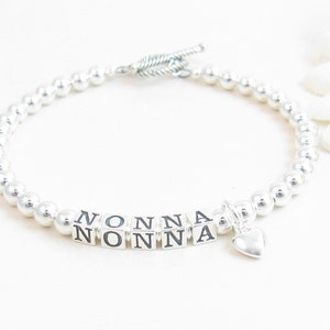 May include: A silver beaded bracelet with the word "NONNA" spelled out in silver letter beads. A small silver heart charm dangles from the bracelet. The bracelet has a toggle clasp.