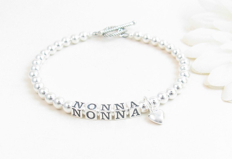 May include: A silver beaded bracelet with the word "NONNA" spelled out in silver letter beads. A small silver heart charm dangles from the bracelet. The bracelet has a toggle clasp.