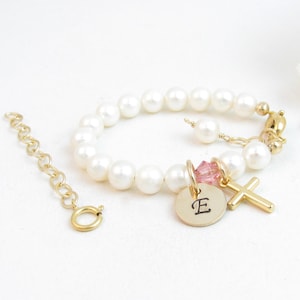 May include: A gold pearl bracelet with a small cross charm, a pink crystal charm, and a gold disc charm with the letter "E" engraved on it.