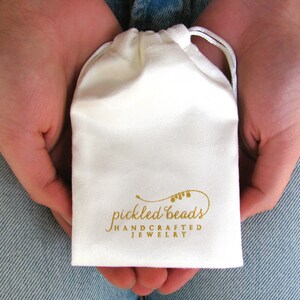 May include: A white drawstring pouch with the text "pickled beads handcrafted jewelry" printed in gold.