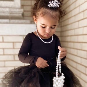May include: A young girl wearing a black tulle skirt and a black long-sleeved top with a pearl necklace. She is holding a white pearl handbag and has a sparkly tiara in her hair.