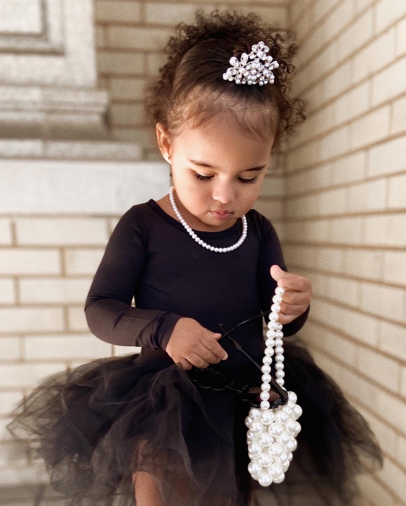 May include: A young girl wearing a black tulle skirt and a black long-sleeved top with a pearl necklace. She is holding a white pearl handbag and has a sparkly tiara in her hair.