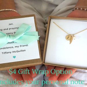 May include: A gold necklace with a small round charm, a feather charm, and a pearl charm. The necklace is in a brown gift box with a light blue ribbon and a handwritten note. The note says "Jen. Thinking and praying sorry for much condolence, my friend. Tiffany McQuillen". The box is labelled "&pound;4 Gift Wrap Option Includes your personal note".