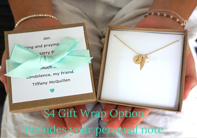 May include: A gold necklace with a small round charm, a feather charm, and a pearl charm. The necklace is in a brown gift box with a light blue ribbon and a handwritten note. The note says "Jen. Thinking and praying sorry for much condolence, my friend. Tiffany McQuillen". The box is labelled "&pound;4 Gift Wrap Option Includes your personal note".
