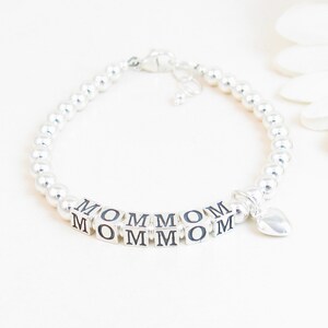 May include: A silver beaded bracelet with the word "MOM" spelled out in silver letter blocks. A small heart charm dangles from the bracelet. The bracelet is on a white background.