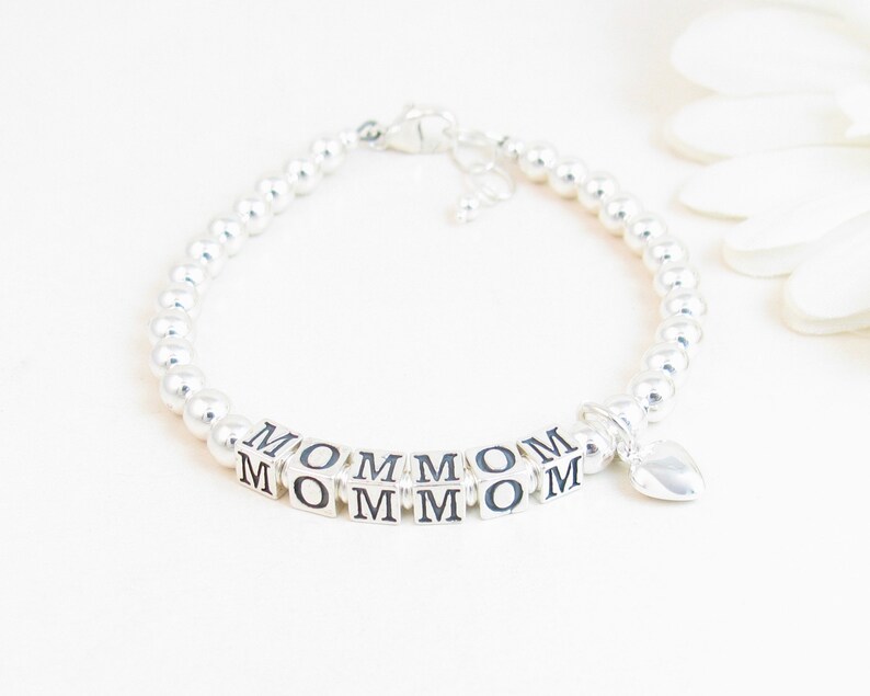 May include: A silver beaded bracelet with the word "MOM" spelled out in silver letter blocks. A small heart charm dangles from the bracelet. The bracelet is on a white background.