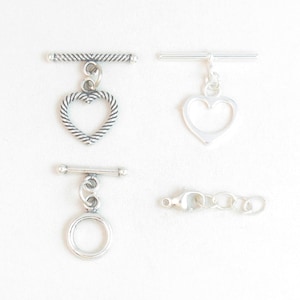 May include: Four silver-toned jewelry clasps. Two are toggle clasps with heart-shaped charms, one has a round charm, and the last is a lobster claw clasp with a chain.