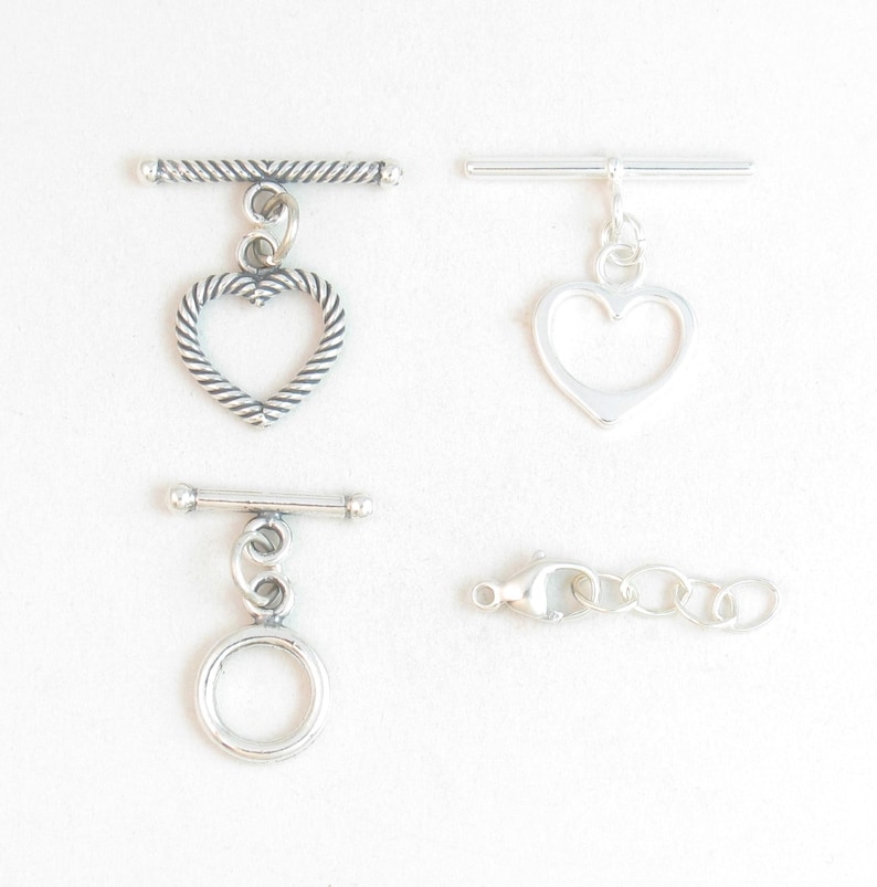 May include: Four silver-toned jewelry clasps. Two are toggle clasps with heart-shaped charms, one has a round charm, and the last is a lobster claw clasp with a chain.