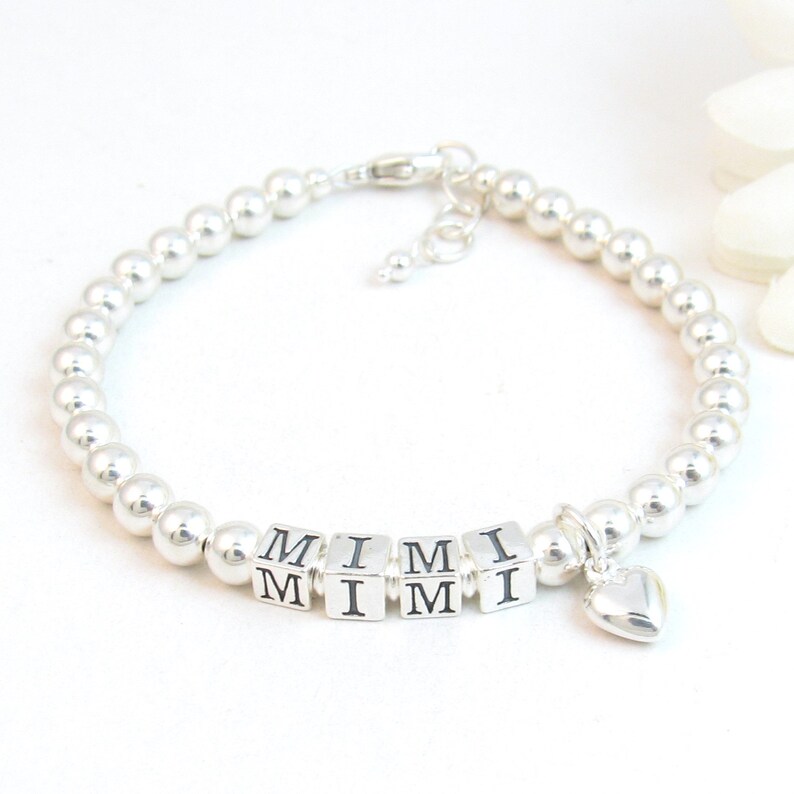 May include: A silver beaded bracelet with the name "MIMI" spelled out in square letter beads. A small silver heart charm dangles from the bracelet. The bracelet is adjustable and has a lobster clasp.