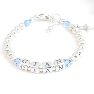 May include: A silver bracelet with the name "CIAN" spelled out in silver lettered cubes. The bracelet features small silver beads and light blue crystal accents. A small cross charm hangs from the bracelet.