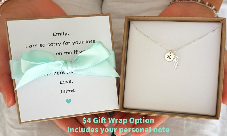 Puede incluir: Una caja de regalo con una cinta azul claro y una nota escrita a mano que dice "Emily, lo siento mucho por tu p&eacute;rdida sobre m&iacute; si estuvieras aqu&iacute;... Con amor, Jaime". La caja contiene un collar de plata con un peque&ntilde;o dije que tiene una letra "K" y un dise&ntilde;o de ala. El texto "4 &euro; Opci&oacute;n de envoltorio de regalo Incluye tu nota personal" est&aacute; impreso en la parte inferior de la caja.
