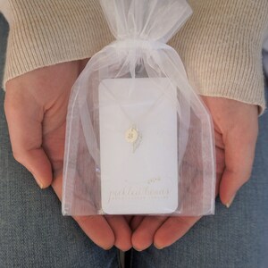 Puede incluir: Una bolsa de organza blanca contiene un delicado collar de plata con un delicado dije de pluma y un dije con la letra 'B'. El collar se presenta en una tarjeta blanca con el texto "pickled beans handmade jewelry".