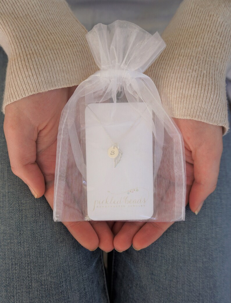 Puede incluir: Una bolsa de organza blanca contiene un delicado collar de plata con un delicado dije de pluma y un dije con la letra 'B'. El collar se presenta en una tarjeta blanca con el texto "pickled beans handmade jewelry".