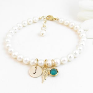 May include: A pearl bracelet with a gold charm featuring a small angel wing and a letter 'J' charm. A green gemstone charm is also attached to the bracelet.