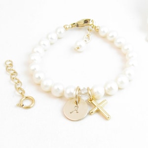 May include: A gold-toned bracelet featuring white spherical beads, a gold-toned lobster clasp, and an extender chain. Two charms hang from the centre: a circular gold-toned disc engraved with the letter A and a simple gold-toned cross charm.