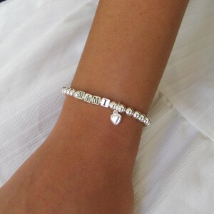 May include: A silver beaded bracelet spelling "MIMI" with a heart charm. The bracelet is on a wrist, with a white fabric background. The bracelet is made of small, round beads.
