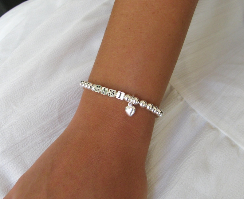 May include: A silver beaded bracelet spelling "MIMI" with a heart charm. The bracelet is on a wrist, with a white fabric background. The bracelet is made of small, round beads.