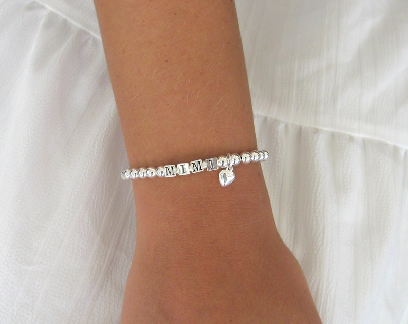 May include: Silver beaded bracelet spelling "MIMI" with a small heart charm. The bracelet is worn on a wrist against a white background. The letters are in a block style.