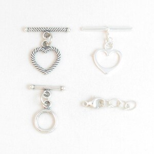 May include: Four silver-toned jewelry clasps and connectors are displayed against a white background. The clasps include a toggle clasp with a textured heart, a smooth heart, and a circle. A lobster clasp with a chain is also shown.