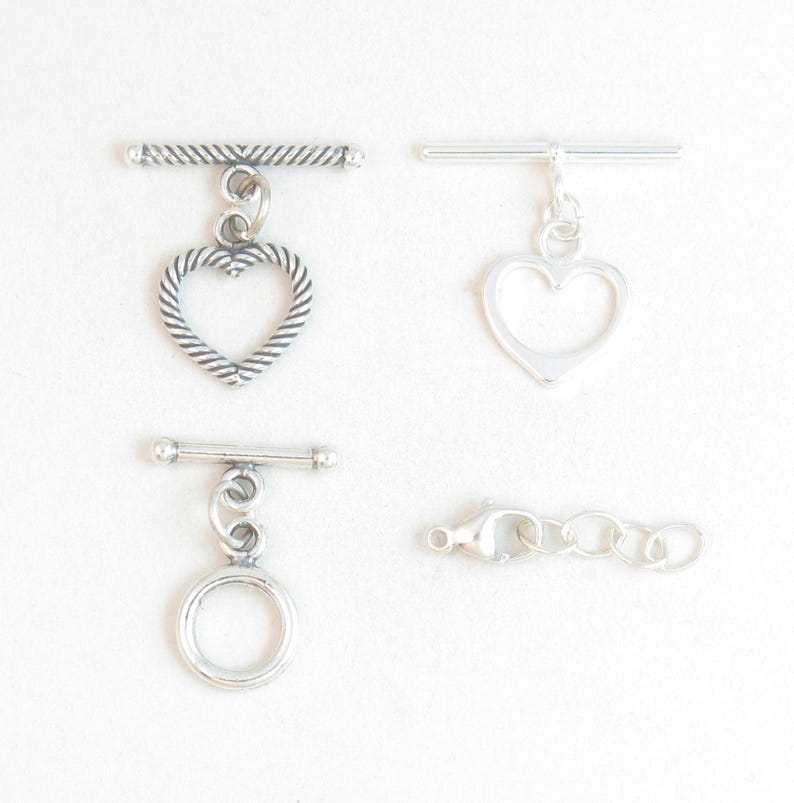 May include: Four silver-toned jewelry clasps and connectors are displayed against a white background. The clasps include a toggle clasp with a textured heart, a smooth heart, and a circle. A lobster clasp with a chain is also shown.