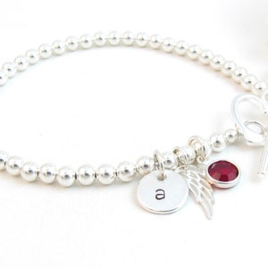 May include: Silver bead bracelet with a heart-shaped clasp, a small charm with the letter 'a', a silver angel wing charm, and a red gemstone charm.