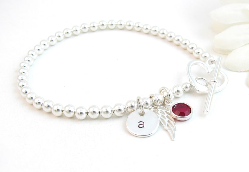 May include: Silver bead bracelet with a heart-shaped clasp, a small charm with the letter 'a', a silver angel wing charm, and a red gemstone charm.