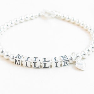 May include: A silver beaded bracelet with the name "MILLIE" spelled out in silver cube beads. A small silver heart charm dangles from the bracelet. The bracelet is on a white background.