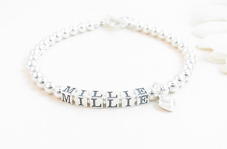 May include: A silver beaded bracelet with the name "MILLIE" spelled out in silver cube beads. A small silver heart charm dangles from the bracelet. The bracelet is on a white background.