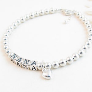 May include: A silver beaded bracelet with the word "NANA" spelled out in square letter beads. A small heart charm dangles from the bracelet. The bracelet is on a white surface.