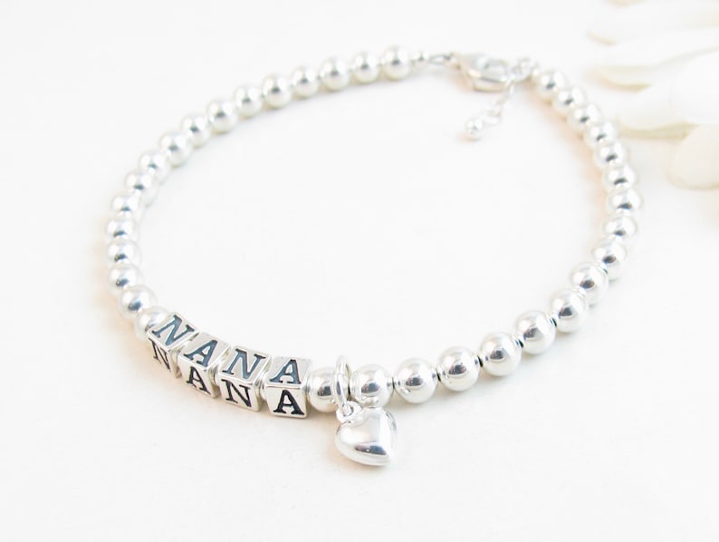 May include: A silver beaded bracelet with the word "NANA" spelled out in square letter beads. A small heart charm dangles from the bracelet. The bracelet is on a white surface.