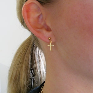 May include: A gold cross earring with a small ball at the top. The earring is worn in the left ear of a person with blonde hair.