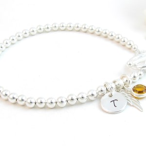 May include: Silver bead bracelet with a charm that has the letter "T" on it, a small amber gemstone, and a silver angel wing charm.